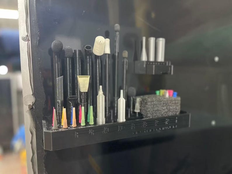 May include: A black plastic organizer shelf mounted on a dark surface, featuring multiple vertical slots holding various black and silver tools, small colorful nozzles, and a textured gray foam block for storing additional small components.