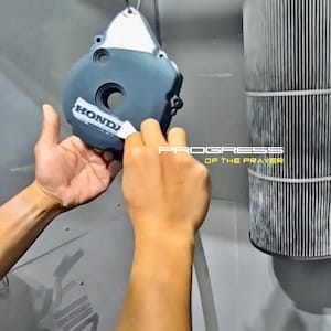 May include: A dark gray metal engine cover featuring a silver Honda logo decal being applied. The circular component has a central opening and is suspended by a hook. The text PROGRESS OF THE PRAYER is visible in the background in white and yellow.