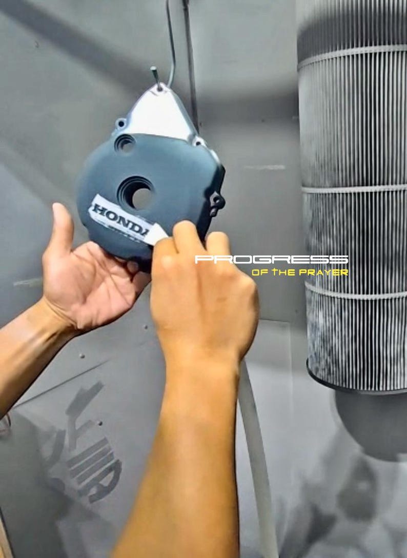 May include: A dark gray metal engine cover featuring a silver Honda logo decal being applied. The circular component has a central opening and is suspended by a hook. The text PROGRESS OF THE PRAYER is visible in the background in white and yellow.