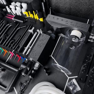 May include: A professional airbrush cleaning station kit featuring a black main unit with a clear cylindrical reservoir, various black metal cleaning tools, assorted colorful plastic nozzle tips, white foam swabs, and a flexible white silicone hose.