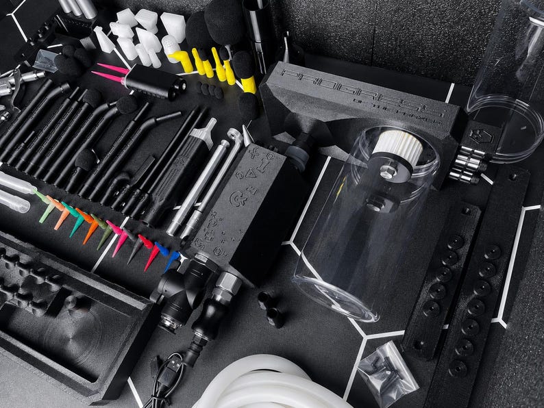May include: A professional airbrush cleaning station kit featuring a black main unit with a clear cylindrical reservoir, various black metal cleaning tools, assorted colorful plastic nozzle tips, white foam swabs, and a flexible white silicone hose.