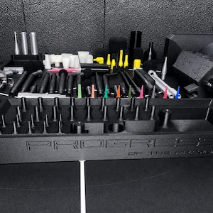 May include: A black rectangular organizer tray with the text PROGRESS OF THE PRINTER embossed on the front. The tray features numerous small vertical pegs and compartments for holding various black, silver, and colorful tool attachments.
