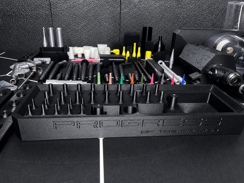 May include: A black rectangular organizer tray with the text PROGRESS OF THE PRINTER embossed on the front. The tray features numerous small vertical pegs and compartments for holding various black, silver, and colorful tool attachments.
