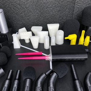 May include: A collection of audio equipment accessories including black foam microphone covers, white plastic attachments in various shapes, yellow nozzles, two bright pink pointed tools, and a black rectangular power bank with a textured finish.
