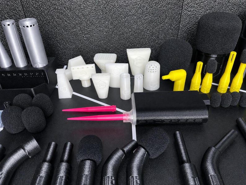 May include: A collection of audio equipment accessories including black foam microphone covers, white plastic attachments in various shapes, yellow nozzles, two bright pink pointed tools, and a black rectangular power bank with a textured finish.