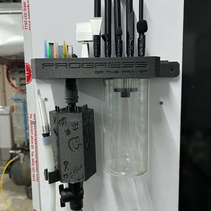 May include: A black 3D-printed wall-mounted organizer labeled PROGRESS OF THE PRAYER, featuring slots for various black and white tools, a clear cylindrical container, and a black hexagonal block embossed with letters and symbols attached to tubing.