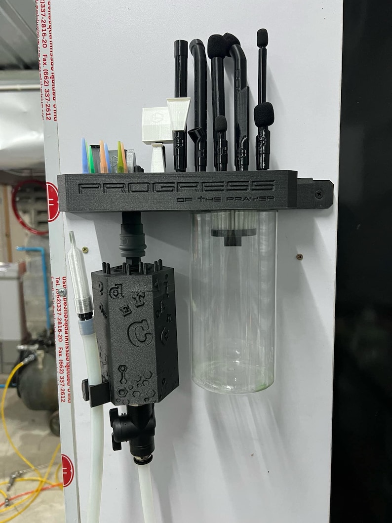 May include: A black 3D-printed wall-mounted organizer labeled PROGRESS OF THE PRAYER, featuring slots for various black and white tools, a clear cylindrical container, and a black hexagonal block embossed with letters and symbols attached to tubing.