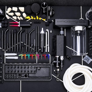 Op de afbeelding: Een uitgebreide set airbrush-apparatuur in zwart en zilver, inclusief een hoofdeenheid met het label PROGRESS OF THE MYTH, een doorzichtige glazen container, een opgerolde witte slang, diverse zwarte mondstukken, kleurrijke tips en een zilveren luchtpistool.