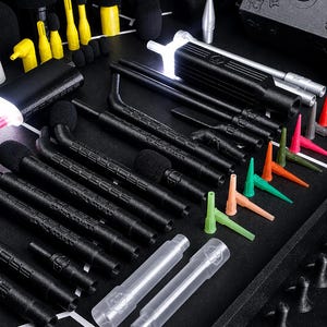 May include: A collection of black, textured, cylindrical tools with integrated LED lights, accompanied by various small, colorful, pointed plastic nozzles in red, blue, green, orange, and yellow, arranged on a dark, gridded surface.