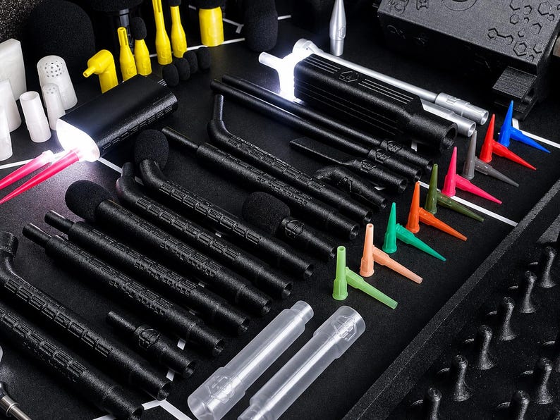 May include: A collection of black, textured, cylindrical tools with integrated LED lights, accompanied by various small, colorful, pointed plastic nozzles in red, blue, green, orange, and yellow, arranged on a dark, gridded surface.