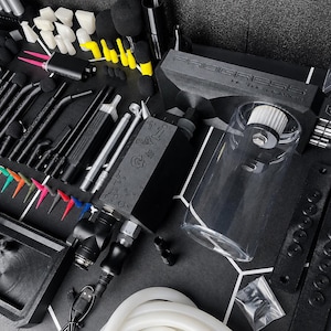 May include: A professional airbrush kit featuring a black air compressor with the text PROGRESS, a clear cylindrical reservoir, various black airbrush pens, assorted colorful needle tips, black foam swabs, and a white silicone hose on a black mat.