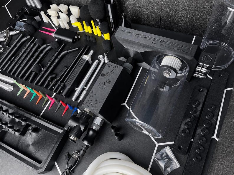 May include: A professional airbrush kit featuring a black air compressor with the text PROGRESS, a clear cylindrical reservoir, various black airbrush pens, assorted colorful needle tips, black foam swabs, and a white silicone hose on a black mat.