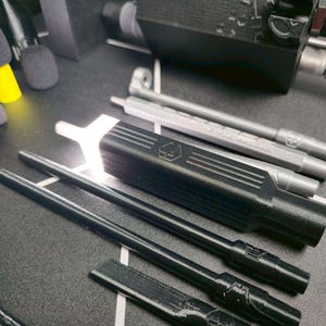 May include: A collection of 3D printed components including a black rectangular housing with a hexagonal logo, several long black and silver cylindrical rods, small black foam microphone covers, and various colorful plastic tips in a set.
