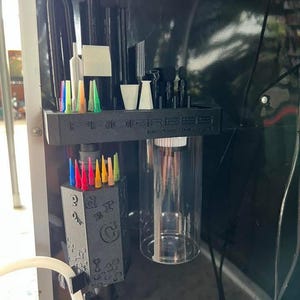 May include: A black 3D-printed organizer shelf labeled PROGRESS, featuring slots for various black tools and colorful needle tips. Below, a matching black hexagonal block holds more colorful needle tips, alongside a clear cylindrical container.