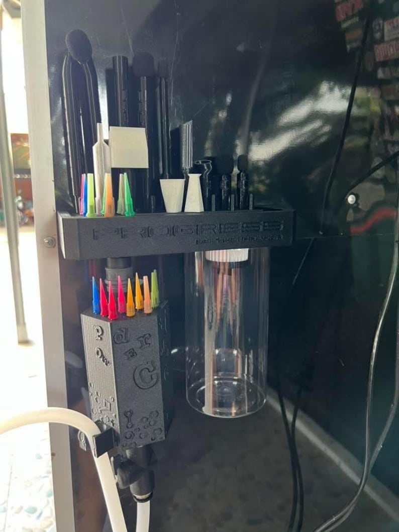 May include: A black 3D-printed organizer shelf labeled PROGRESS, featuring slots for various black tools and colorful needle tips. Below, a matching black hexagonal block holds more colorful needle tips, alongside a clear cylindrical container.