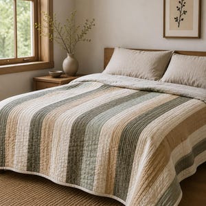 May include: A quilted bedspread featuring vertical stripes in muted shades of sage green, cream, and tan. The fabric has a textured, stitched pattern throughout, covering a bed with two matching pillows in a solid light beige color.
