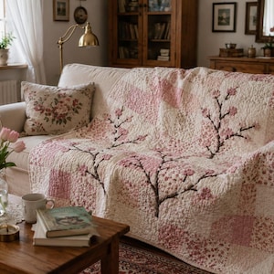 Könnte beinhalten: Ein Patchwork-Quilt in einer cremefarbenen und rosafarbenen Farbpalette mit einem markanten Kirschblütenzweig-Design. Der Quilt besteht aus verschiedenen quadratischen Stoffblöcken mit floralen und einfarbigen Mustern und ist mit detaillierten Steppnähten versehen.
