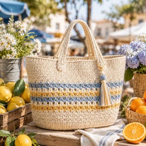 Könnte beinhalten: Eine beige Häkeltasche mit horizontalen Streifen in Hellblau und Senfgelb. Die Tasche verfügt über zwei stabile Tragegriffe und eine dekorative Quaste an einer Seite, die aus hellblauen, senfgelben und beigen Garnfäden besteht.