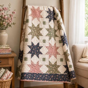 May include: A cream-coloured quilt with a star pattern in shades of pink, green, and navy blue. The quilt is draped over a wooden ladder, showcasing its intricate design and floral accents. The quilt's border features a navy blue floral pattern.