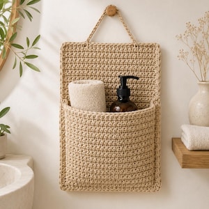 Könnte beinhalten: Ein beigefarbener, gehäkelter Wand-Organizer mit einer rechteckigen Rückwand und einer tiefen Vordertasche. In der Tasche befinden sich ein gerolltes beiges Handtuch und eine dunkelbraune Kunststoff-Pumpflasche. Aufgehängt an einer passenden Kordel.