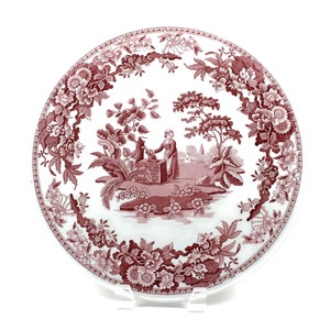 May include: A white ceramic plate with a red floral pattern and a central image of a woman drawing water from a well. The plate has a scalloped edge.