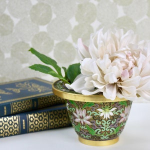 May include: A white flower with pink accents sits in a small, green and gold cloisonné vase with floral designs. The vase is sitting on top of a stack of three blue and gold books.