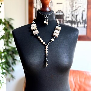 May include: A necklace and earring set featuring rectangular patterned beads in shades of beige and brown, interspersed with matte black spherical beads and smaller clear faceted beads. The necklace has a Y-shaped drop design with a central pendant.