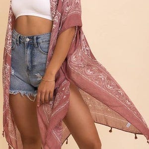 May include: A dusty rose kimono featuring a white paisley print, open front, and short sleeves. The garment is finished with small tassels along the bottom hem, providing a lightweight, flowing silhouette that drapes down to the mid-thigh area.