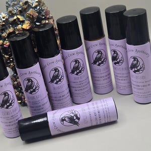 May include: An assortment of roll-on essential oil blends from Violet Crow Apothecary. The lavender-colored labels feature a crow illustration and product names like "Citrus Aura" and "Deep Rest". Black caps top the glass bottles.