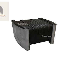 Op de afbeelding: Zwarte leren poef met een geribbelde bovenkant en gebogen armleuningen. De zijkanten zijn gemaakt van geklonken, geborsteld aluminium. De poef heeft een laag profiel en een modern, industrieel ontwerp. De merknaam "Rumaglow" is zichtbaar.