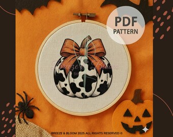 May include: A cross-stitch pattern featuring a pumpkin with a cow print design and an orange bow. The design is set in a wooden embroidery hoop. The background is orange with Halloween-themed decorations.