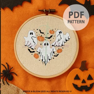 May include: A Halloween-themed embroidery hoop featuring three white ghosts, orange pumpkins, and black bats arranged in a heart shape. The design is on a beige fabric, set in a wooden hoop, with a PDF pattern label. The background is orange.
