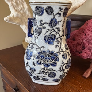 May include: White and blue ceramic vase with a floral design. The vase features a detailed pattern of blue flowers and leaves against a white background. It has two blue handles and a decorative rim.