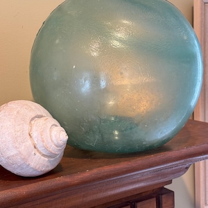 May include: A large, round, seafoam green glass float with a textured surface sits next to a white seashell on a dark wooden shelf. The float is a sphere, and the seashell has a spiral pattern.