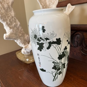May include: White ceramic vase with a floral design in black and green. The vase features an Asian-inspired illustration of flowers and foliage. The vase has a rounded body and a slightly flared rim. The vase has text in the image.