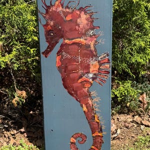 May include: A hand-painted seahorse on a weathered blue wooden plank. The seahorse is primarily shades of brown and orange, with white accents. The seahorse's tail is curled, and the background is a muted blue. The artwork is displayed outdoors.
