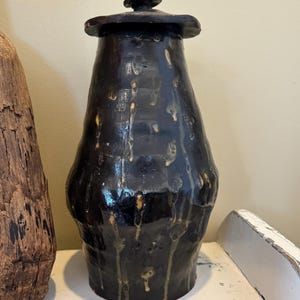 May include: A black ceramic jar with a lid. The jar has a bulbous shape with a narrow neck and a lid. The surface has a glossy finish with drips of a lighter color. The jar is sitting on a white surface.