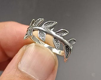 May include: A silver-colored ring featuring a delicate leaf design. The ring showcases a series of detailed leaves, creating a nature-inspired aesthetic. The ring is held up against a neutral background, highlighting the intricate details of the jewelry.