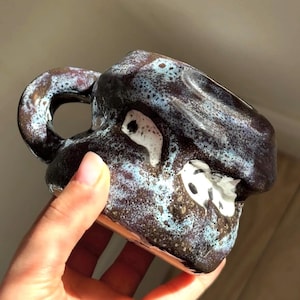 Puede incluir: Una taza de cerámica hecha a mano con una forma orgánica e irregular. La superficie presenta un esmalte marrón oscuro moteado con parches de color azul claro y blanco, creando un aspecto texturizado. Tiene una sola asa y un acabado brillante.