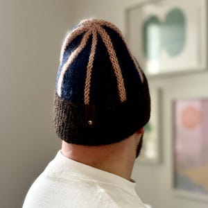 May include: A knitted beanie hat with a color block design. The hat features a dark blue base with vertical tan stripes and a tan top. The bottom portion of the hat is brown. A small brown leather tag is sewn on the side.