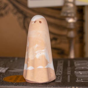May include: A ceramic ghost figurine with a peach-colored body and white accents. The ghost has two small eye holes and a rounded top. The figurine is a decorative item, likely for Halloween or spooky-themed decor.