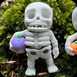 Könnte beinhalten: Eine hellgraue, 3D-gedruckte Skelettfigur mit großem Kopf und hohlen Augenhöhlen. Das Skelett hat einen Brustkorb sowie ein Becken und hält in der rechten Hand einen kleinen, runden, violetten Gegenstand mit einem grünen Stiel.
