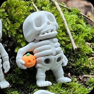 Könnte beinhalten: Eine kleine, weiße, stilisierte Skelettfigur mit großem Kopf und abgerundeten Zügen. Das Skelett hält einen winzigen, runden, orangefarbenen Kürbis mit einem grünen Stiel in der rechten Hand. Die Figur hat eine glatte, matte Oberfläche.