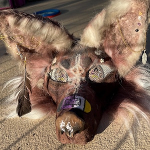 May include: A brown and white faux fur animal mask featuring large pointed ears adorned with gold chains, small crystals, and a hanging feather. The mask has mesh eye coverings, a glossy brown nose, and a rectangular patch with purple and yellow.