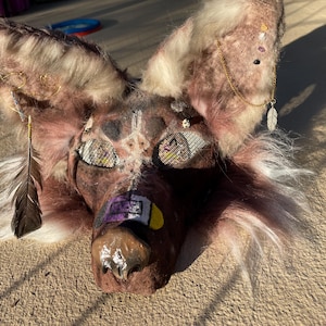 May include: A handmade, brown and white furry animal mask with detailed features. The mask has mesh eyes, a painted nose, and decorative feathers and jewelry. The ears are adorned with gold wire and small embellishments. The overall design is intricate and artistic.