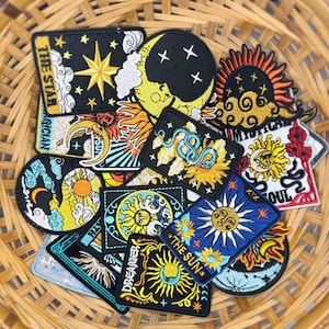May include: A selection of embroidered patches with celestial and mystical themes. The patches depict the sun, moon, stars, and snakes, with text including "The Star" and "The Sun". The patches are displayed in a woven basket.