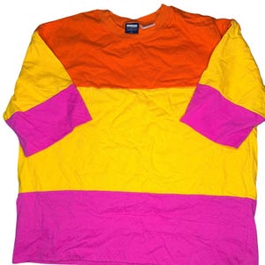 May include: A short-sleeve t-shirt featuring a colour-blocked design with a horizontal orange upper section, a yellow middle section, and a pink lower section. The sleeves are yellow at the top and pink at the bottom. A small dark blue tag is at the neck.
