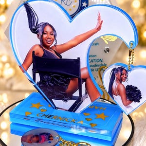 May include: A blue heart-shaped acrylic display featuring a photograph of a person in a black dress, a smaller heart-shaped keychain with a photo, and a circular keychain. The base is blue with gold stars and the text CHERNICKA printed on it.