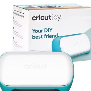 May include: A teal and white Cricut Joy cutting machine with a rounded rectangular shape. The machine is positioned in front of its product packaging box, which features the text 'cricut joy' and 'Your DIY best friend' in dark blue lettering.