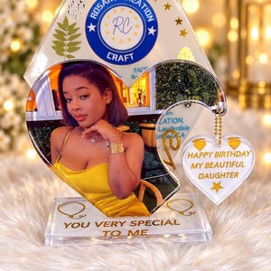 May include: A clear acrylic heart-shaped photo plaque featuring a portrait of a person in a yellow dress. A smaller heart charm hangs from the side with the text HAPPY BIRTHDAY MY BEAUTIFUL DAUGHTER. The base reads YOU VERY SPECIAL TO ME.